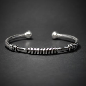 May include: A silver cuff bracelet with a central band featuring a spiral design. The bracelet has rounded ball-shaped ends. The jewellery is shown against a dark background.