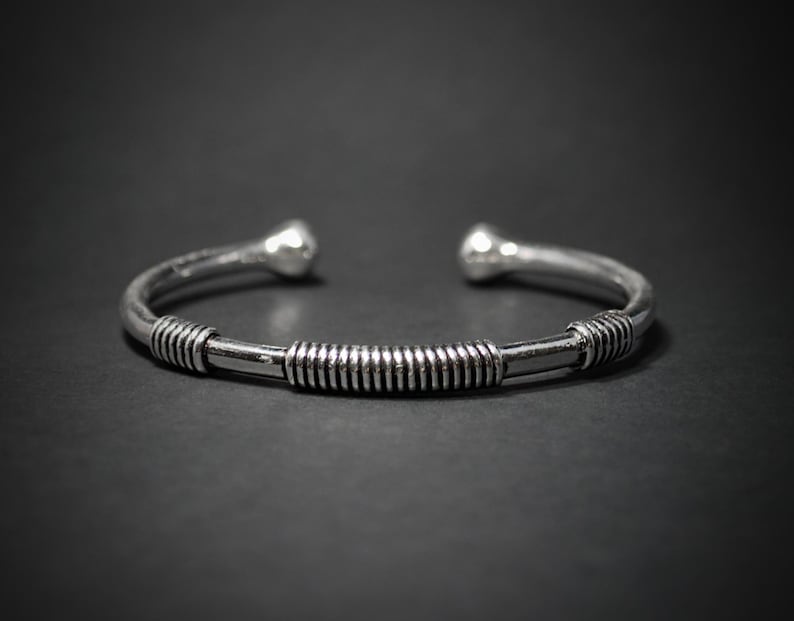 May include: A silver cuff bracelet with a central band featuring a spiral design. The bracelet has rounded ball-shaped ends. The jewellery is shown against a dark background.