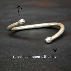 May include: A silver-coloured, open cuff bracelet with rounded ball ends. The bracelet is curved and has a gap for easy wear. The text "To put it on, open it like this" is at the bottom of the image.