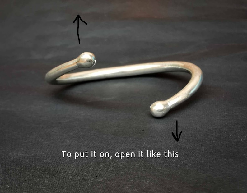 May include: A silver-coloured, open cuff bracelet with rounded ball ends. The bracelet is curved and has a gap for easy wear. The text "To put it on, open it like this" is at the bottom of the image.