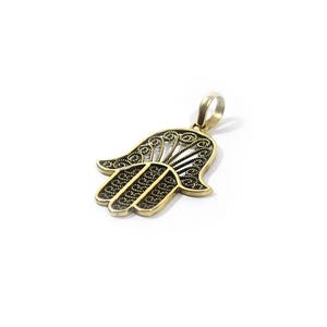 May include: A gold-coloured Hamsa hand pendant with detailed filigree work. The pendant has a stylised hand shape with openwork patterns and a loop for a chain. The design is set against a white background.