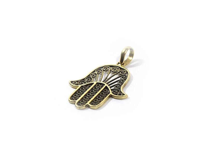 May include: A gold-coloured Hamsa hand pendant with detailed filigree work. The pendant has a stylised hand shape with openwork patterns and a loop for a chain. The design is set against a white background.