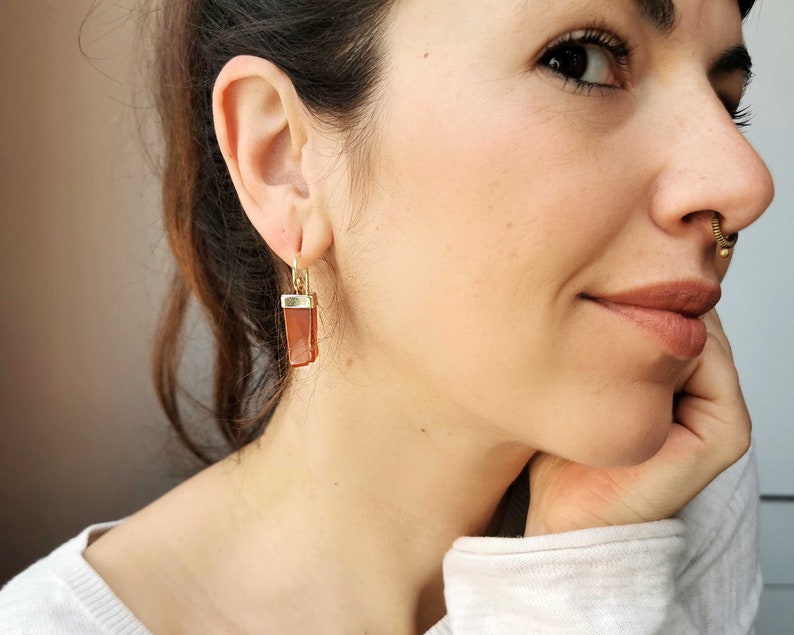 Puede incluir: Un par de pendientes de aro de oro con un colgante de piedra de cornalina marr&oacute;n. Los pendientes cuelgan del l&oacute;bulo de la oreja de una persona.
