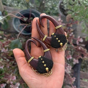 May include: A pair of dark brown wooden ear weights shaped like hooks, featuring black macrame cord detailing with small gold-toned beads and thin gold-toned wire accents. Each piece includes a delicate gold-toned chain draped along the bottom.