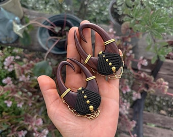 May include: A pair of dark brown wooden ear weights shaped like hooks, featuring black macrame cord detailing with small gold-toned beads and thin gold-toned wire accents. Each piece includes a delicate gold-toned chain draped along the bottom.