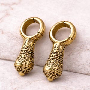 May include: Two ornate, gold-colored ear weights. Each weight features a circular top and a decorative, teardrop-shaped bottom with detailed patterns. The weights are designed with a split ring for easy insertion and removal. The background is a light, textured surface.