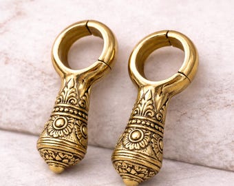 May include: Two ornate, gold-colored ear weights. Each weight features a circular top and a decorative, teardrop-shaped bottom with detailed patterns. The weights are designed with a split ring for easy insertion and removal. The background is a light, textured surface.