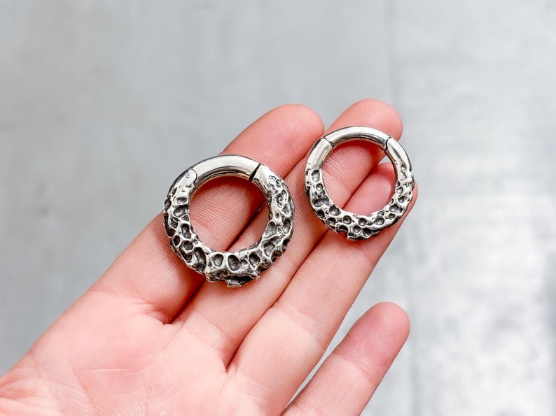 May include: Two silver-colored, textured hoop earrings. The earrings have a unique, organic design with a rough, pitted surface. The hoops are of different sizes, with a hinged clasp for closure. Jewelry.