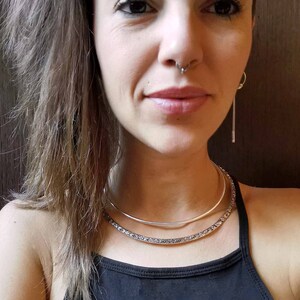 May include: A woman wearing a black tank top with two silver necklaces. The necklaces are layered, with a thin, smooth choker on top of a thicker, textured choker. The woman has long brown hair and is wearing a nose ring.