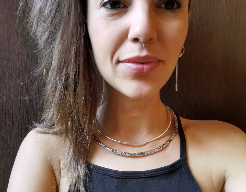 May include: A woman wearing a black tank top with two silver necklaces. The necklaces are layered, with a thin, smooth choker on top of a thicker, textured choker. The woman has long brown hair and is wearing a nose ring.