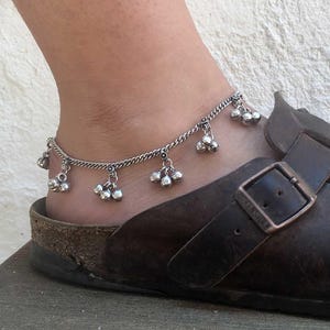 Op de afbeelding: Een zilveren enkelband met bungelende belletjes. De enkelband wordt gedragen op een enkel, gecombineerd met een bruine leren sandaal met een gesp.