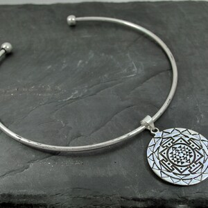 May include: A silver choker necklace with a round pendant featuring a geometric design. The pendant is detailed with a mandala-like pattern.