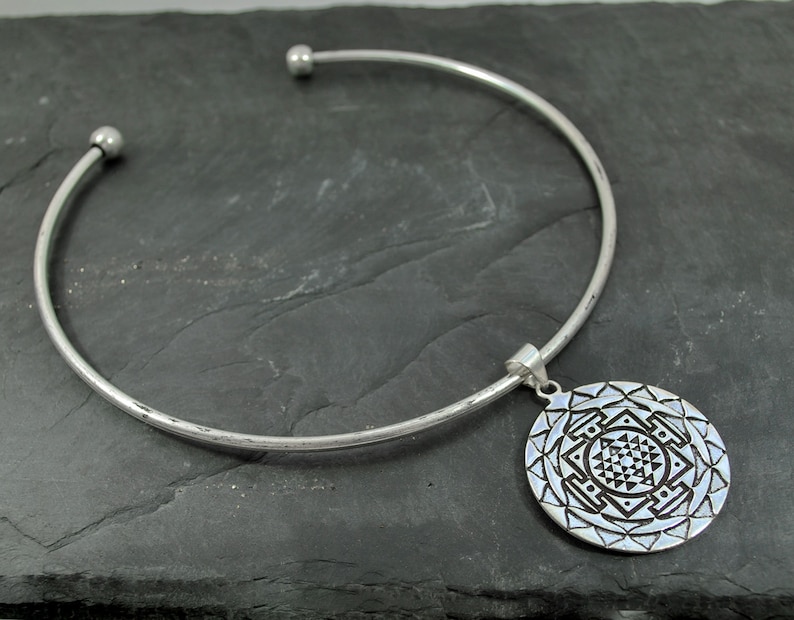 May include: A silver choker necklace with a round pendant featuring a geometric design. The pendant is detailed with a mandala-like pattern.