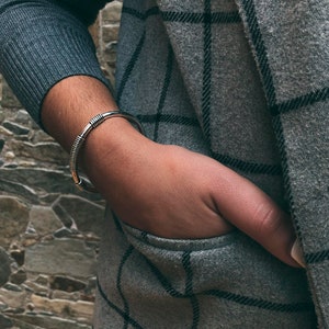 May include: A silver cuff bracelet with a coiled design. The bracelet is worn on a wrist, with a grey sweater and a grey plaid coat in the background. The person's hand is in the coat pocket.