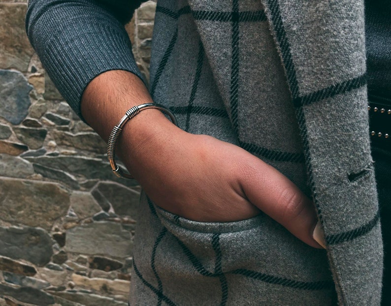May include: A silver cuff bracelet with a coiled design. The bracelet is worn on a wrist, with a grey sweater and a grey plaid coat in the background. The person's hand is in the coat pocket.