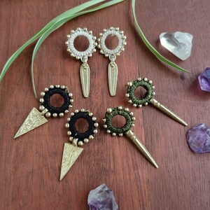 Może przedstawiać: Ręcznie robione kolczyki w kolorach białym, czarnym i oliwkowym. Każdy kolczyk ma okrągły wzór z centralnym pierścieniem i mosiężną, przypominającą strzałę zawieszką. Kolczyki są prezentowane na drewnianej powierzchni.