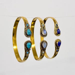 Può includere: Tre braccialetti d'oro con pietre preziose di diversi colori. I braccialetti sono impilati uno sopra l'altro. Le pietre preziose sono turchese, pietra di luna e lapislazzuli.