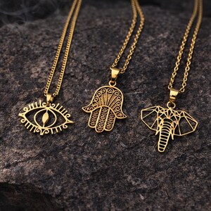 May include: Three gold-coloured pendant necklaces. The pendants feature an eye, a Hamsa hand, and an elephant. Each necklace has a delicate chain. The necklaces are displayed on a dark, textured surface.