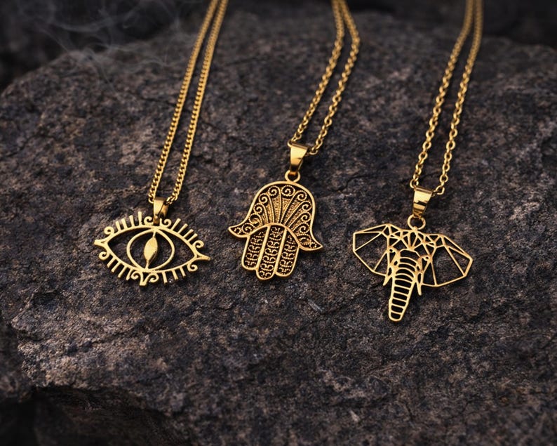 May include: Three gold-coloured pendant necklaces. The pendants feature an eye, a Hamsa hand, and an elephant. Each necklace has a delicate chain. The necklaces are displayed on a dark, textured surface.