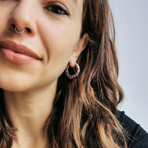 May include: A close-up shot of a person wearing a silver septum ring and a silver hoop earring. The earring has a decorative pattern around the hoop. The person has long, wavy brown hair and is wearing a black top.