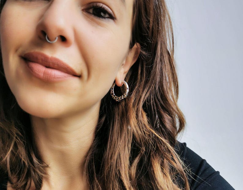 May include: A close-up shot of a person wearing a silver septum ring and a silver hoop earring. The earring has a decorative pattern around the hoop. The person has long, wavy brown hair and is wearing a black top.