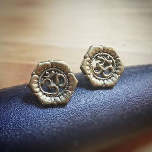 Op de afbeelding: Een paar gouden oorbellen met een lotusbloem ontwerp met een Om symbool in het midden.