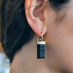 Puede incluir: Pendientes de piedra negra con engaste dorado. Los pendientes cuelgan de la oreja de una persona.