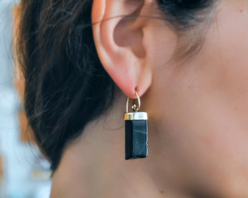 Puede incluir: Pendientes de piedra negra con engaste dorado. Los pendientes cuelgan de la oreja de una persona.