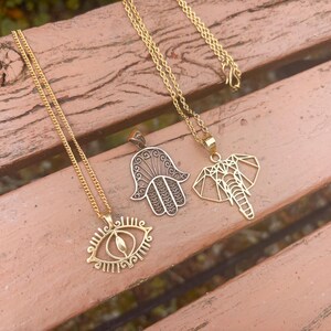 May include: Three gold-coloured necklaces with different pendant designs are displayed on a wooden surface. One features an eye, another a hand, and the third an elephant. The chains are delicate, and the pendants are intricately detailed.