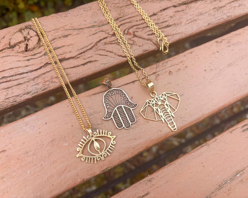 May include: Three gold-coloured necklaces with different pendant designs are displayed on a wooden surface. One features an eye, another a hand, and the third an elephant. The chains are delicate, and the pendants are intricately detailed.