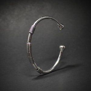 May include: Silver cuff bracelet with a minimalist design. The open cuff has a rounded end and a wrapped section with parallel lines. The bracelet is set against a dark background.