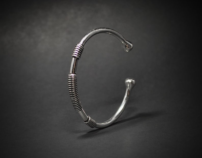 May include: Silver cuff bracelet with a minimalist design. The open cuff has a rounded end and a wrapped section with parallel lines. The bracelet is set against a dark background.