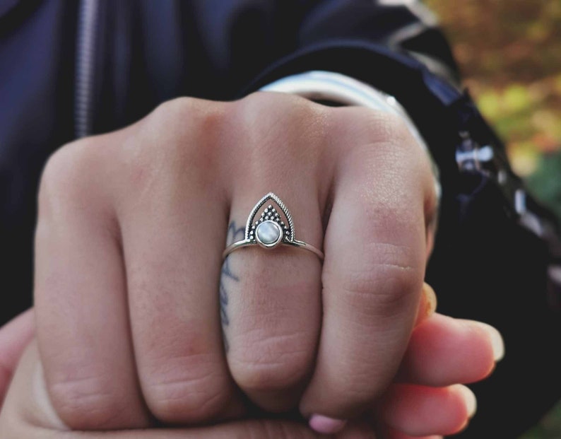 May include: A silver ring with a white stone set in a geometric design. The ring is on a person's finger.