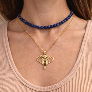 May include: A gold elephant pendant necklace with a geometric design, layered with a dark blue beaded choker necklace. The necklaces are displayed on a person wearing a light tan vest top.