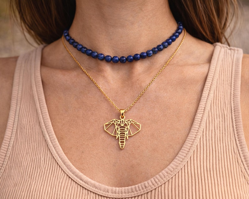 May include: A gold elephant pendant necklace with a geometric design, layered with a dark blue beaded choker necklace. The necklaces are displayed on a person wearing a light tan vest top.