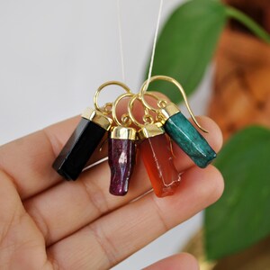 Puede incluir: Cinco pendientes de tono dorado con gotas de piedras preciosas de diferentes colores. Los pendientes presentan piedras preciosas negras, rojas, naranjas y verdes.