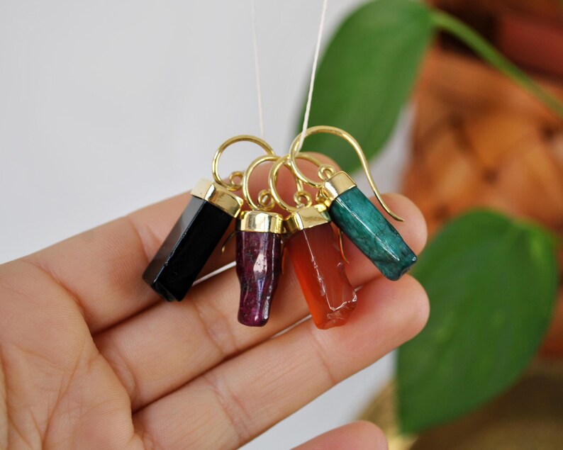 Puede incluir: Cinco pendientes de tono dorado con gotas de piedras preciosas de diferentes colores. Los pendientes presentan piedras preciosas negras, rojas, naranjas y verdes.