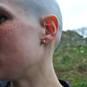 May include: Close-up of a person's face with a shaved head and a small stud earring in their left ear.