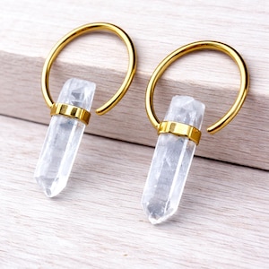 May include: A pair of gold-colored hoop earrings with clear quartz crystal pendants. Each earring features a polished crystal suspended from a gold hoop, with a gold band securing the crystal. The earrings are displayed on a light-colored wooden surface.