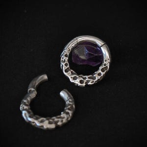 May include: Two silver-colored, textured hoop earrings. One earring is open, while the other is closed and features a purple gemstone. The earrings are set against a black background, highlighting their intricate design and color.