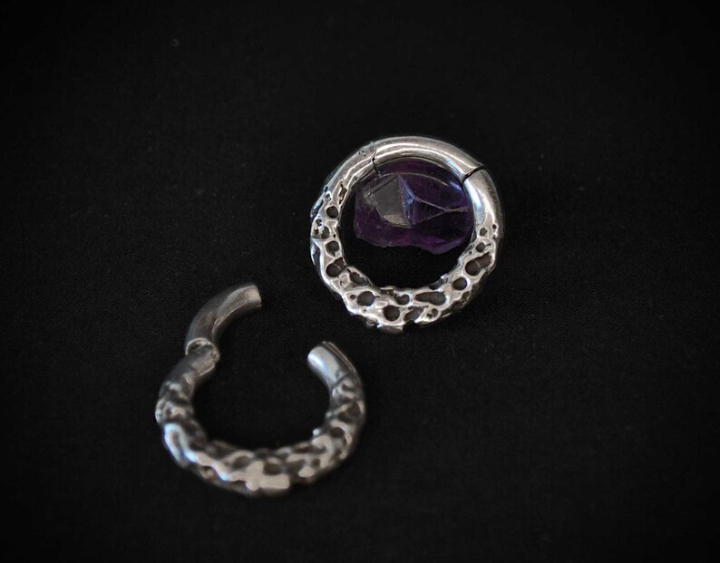 May include: Two silver-colored, textured hoop earrings. One earring is open, while the other is closed and features a purple gemstone. The earrings are set against a black background, highlighting their intricate design and color.