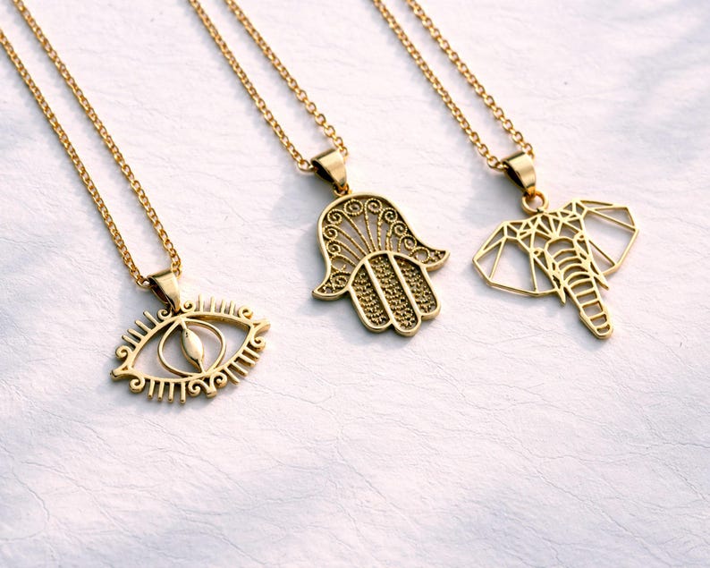 May include: Three gold-coloured pendant necklaces. The pendants feature an eye, a Hamsa hand, and a geometric elephant. Each necklace has a delicate chain. The jewellery is displayed on a white surface.