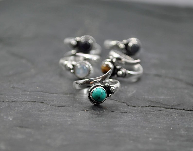 Op de afbeelding: Een set zilveren ringen met verschillende gekleurde edelstenen. De ringen hebben een uniek ontwerp met een gebogen band en een klein, puntig accent. De edelstenen zijn turquoise, maansteen, tijgeroog en zwarte onyx.