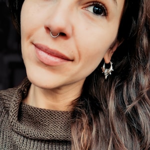 Peut inclure: Une femme aux longs cheveux bruns portant un pull marron. Elle porte un anneau de nez argent&eacute; et une boucle d'oreille argent&eacute;e avec un motif d&eacute;coratif.