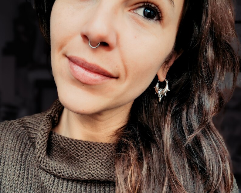 Peut inclure: Une femme aux longs cheveux bruns portant un pull marron. Elle porte un anneau de nez argent&eacute; et une boucle d'oreille argent&eacute;e avec un motif d&eacute;coratif.