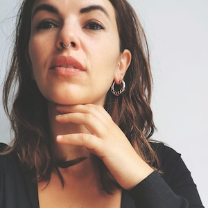 May include: Silver hoop earrings with a textured design are worn by a person with brown hair. A silver nose ring is also visible. The person is wearing a black top and a black choker necklace.