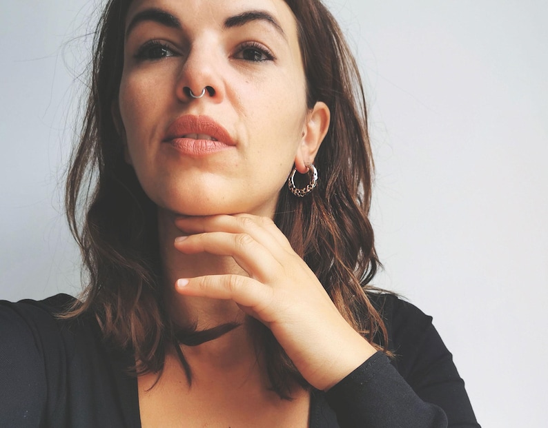 May include: Silver hoop earrings with a textured design are worn by a person with brown hair. A silver nose ring is also visible. The person is wearing a black top and a black choker necklace.