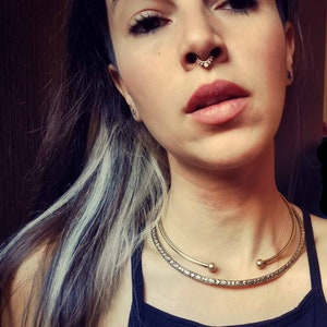 May include: A woman with dark hair and light streaks is wearing a gold choker necklace with a delicate design. She has a septum piercing with a small gold ring.