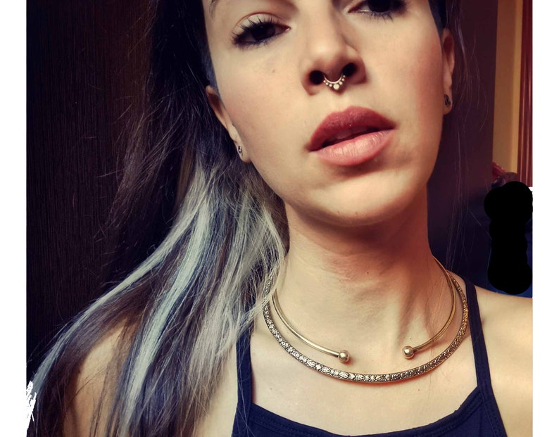 May include: A woman with dark hair and light streaks is wearing a gold choker necklace with a delicate design. She has a septum piercing with a small gold ring.