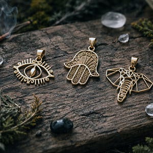 May include: Three gold-coloured pendants with detailed designs are displayed on a weathered wooden surface. The pendants feature an eye, a Hamsa hand, and an elephant. The background includes crystals, greenery, and a lit incense stick, creating a mystical aesthetic.